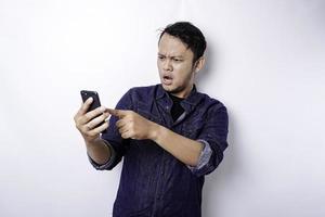 homme asiatique choqué portant une chemise bleue et tenant son téléphone, isolé sur fond blanc photo