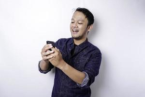 homme asiatique excité portant une chemise bleue souriant tout en tenant son téléphone, isolé sur fond blanc photo