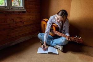 jeune femme hipster assise sur le sol et jouant de la guitare à la maison. adolescente apprenant à jouer de la chanson et à écrire de la musique dans sa chambre. passe-temps, mode de vie, détente, instrument, loisirs, concept d'éducation. photo