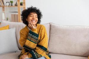 belle fille afro-américaine avec une coiffure afro souriante assise sur un canapé à la maison à l'intérieur. jeune femme africaine aux cheveux bouclés en riant. liberté bonheur insouciant concept de gens heureux. photo