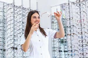 séduisante jeune femme médecin en clinique d'ophtalmologie. le médecin ophtalmologiste se tient près des étagères avec différentes lunettes. jeune médecin dans un magasin d'optique testant des lunettes photo