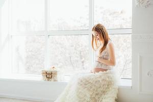 jeune fille enceinte rousse dans une robe blanche près de la fenêtre photo