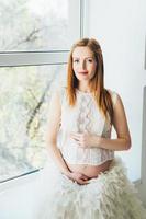 jeune fille enceinte rousse dans une robe blanche près de la fenêtre photo