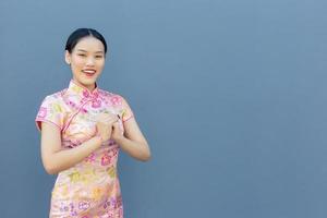 belle femme asiatique aux cheveux longs qui porte une robe cheongsam rose sur le thème du nouvel an chinois alors qu'elle montre sa main comme salutation chinoise avec sur fond gris. photo