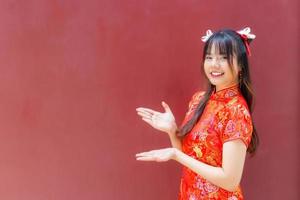 jolie fille asiatique aux cheveux longs qui porte une robe cheongsam rouge sur le thème du nouvel an chinois alors qu'elle montre sa main pour présenter quelque chose sur fond rouge. photo