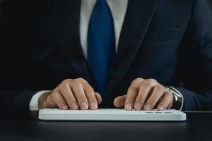 fermer. homme d'affaires de la main portant un costume tapant et travaillant sur un ordinateur à clavier sur une table en bois au bureau à domicile. homme entrepreneur travaillant pour les entreprises au bureau à domicile. concept de technologie de travail. photo