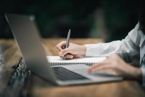 mains en gros plan d'une femme d'affaires travaillant sur un ordinateur portable et utilisant un stylo faisant des avis dans son cahier sur une table en bois avec une tasse de café. notion d'entreprise photo