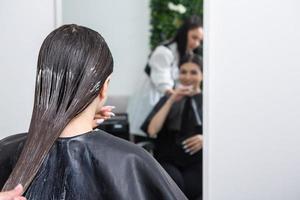 le coiffeur applique un masque capillaire sur les cheveux noirs raides. soins capillaires au salon de beauté. photo