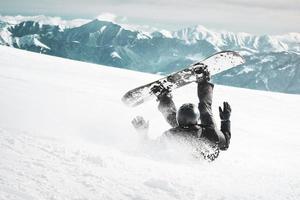 le snowboarder tombe sur le dos avec des éclaboussures de neige sur une piste de ski hors piste enneigée et un vieux télésiège à l'arrière-plan. montagnes du caucase en journée d'hiver ensoleillée. image tonique en noir et blanc. photo