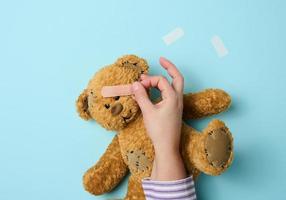 la main féminine tient un ours en peluche brun et colle un pansement adhésif médical sur fond bleu, traitement de tram photo