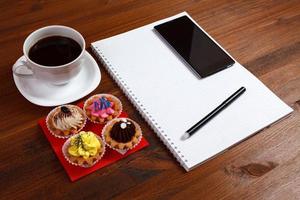 tasse à café blanche avec carnet, quatre cupcakes, smartphone et stylo sur une table en bois. photo