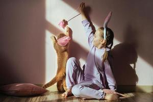 une écolière sur la tête un bandeau avec des oreilles de lapin joue avec un chat domestique rouge dans un chapeau de pâques au soleil photo