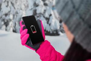 femme tenant un smartphone avec une icône de batterie faible sur l'écran. femme avec chapeau, veste et gants. hiver, temps froid. neige et arbres en arrière-plan photo