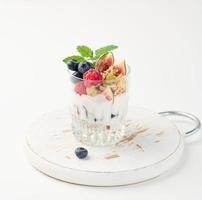 verre transparent avec granola versé avec du yaourt, sur des framboises mûres, des myrtilles et des figues sur un tableau blanc. petit-déjeuner sain photo