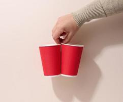 la main tient des tasses en papier carton rouge pour le café, fond beige. vaisselle écologique, zéro déchet photo