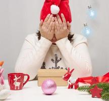 une femme en pull blanc est assise à la table et emballe des cadeaux pour noël. tiens ta tête avec tes mains, tristesse photo