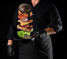 homme adulte dans un uniforme noir tenant une poêle à frire ronde en fonte avec des ingrédients de cheeseburger en lévitation photo