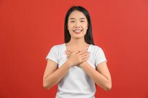 portrait d'une jeune femme asiatique heureuse en chemise bleue montrant son cœur rempli d'amour et se tenant la main sur sa poitrine isolée sur fond rouge photo