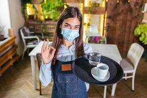 une serveuse dans un masque de protection médicale sert le café au restaurant pendant la pandémie de coronavirus représentant un nouveau concept normal et montrant un signe ok photo