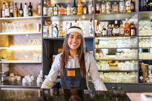 portrait d'un beau barman debout au comptoir souriant et regardant la caméra tout en portant un écran facial en raison de covid-19, des étagères pleines de bouteilles avec de l'alcool en arrière-plan photo