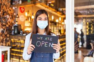 propriétaire d'une petite entreprise avec masque facial tenant le panneau pour la réouverture du lieu après la quarantaine due au covid-19. femme avec masque de protection tenant une pancarte nous sommes ouverts, soutenons les entreprises locales. photo