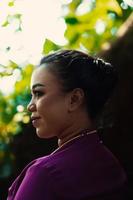 femme indonésienne souriant dans une robe violette traditionnelle tout en portant du maquillage et des bijoux en or devant le grand arbre photo