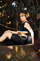 femme indonésienne assise sur le rocher dans un costume de danse noire tout en portant une couronne dorée et un collier doré photo