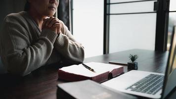 concept de technologie en ligne chrétienne. mains priant de chrétien avec ordinateur portable numérique, église en direct en ligne pour le service du dimanche. une femme catholique asiatique lit le livre de la sainte bible et étudie en ligne. photo