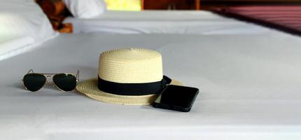 temps de vacances le week-end avec des lunettes de soleil noires, un chapeau de paille vintage blanc et un smartphone mettant sur un lit blanc avec espace de copie. voyage, accessoires, mode et concept de temps de repos photo