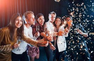 s'amuser avec des cierges magiques. les confettis sont dans l'air. groupe d'amis joyeux célébrant le nouvel an à l'intérieur avec des boissons dans les mains photo