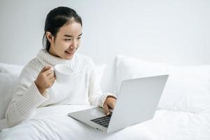 jeune femme jouant sur ordinateur portable et tenant une tasse de café au lit photo
