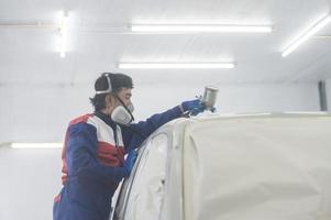 mécanicien automobile asiatique portant une combinaison de protection et un respirateur peint des pare-chocs de carrosserie dans la salle de peinture, pulvérisant des voitures dans la salle de peinture photo