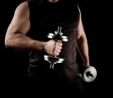 un homme en vêtements noirs tient des haltères en acier dans ses mains, ses muscles sont tendus photo