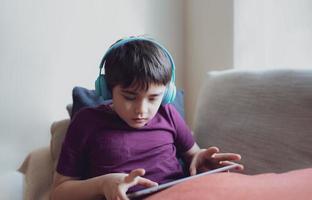 concept d'éducation, portrait clé d'un enfant de l'école primaire lisant un livre électronique sur une tablette pour faire ses devoirs, enfant portant un casque jouant à un jeu en ligne sur internet, garçon assis sur un canapé regardant un dessin animé sur un pavé numérique photo