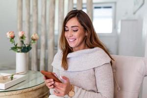 beau message de sa part. jolie jeune femme regardant son téléphone intelligent et souriant assis sur le canapé à la maison. belle femme textos sur son téléphone portable à la maison photo