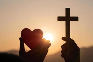 silhouette de mains de femme priant avec croix et tenant une boule de coeur rouge sur fond de lever de soleil nature, crucifix, symbole de la foi. prière de crise de la vie chrétienne à dieu, le concept d'aimer dieu. photo