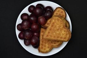 délicieuses gaufres et raisins sur une assiette. un petit-déjeuner sucré composé de gaufres et de raisins rouges. gaufres en forme de cœur. photo