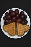délicieuses gaufres et raisins sur une assiette. un petit-déjeuner sucré composé de gaufres et de raisins rouges. gaufres en forme de cœur. photo
