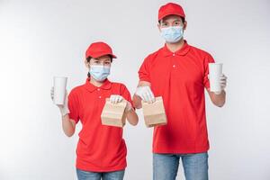 image d'un jeune livreur heureux en casquette rouge t-shirt blanc uniforme masque facial gants debout avec paquet de papier kraft marron vide isolé sur fond gris clair studio photo