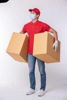 image d'un jeune livreur conscient en casquette rouge t-shirt blanc uniforme masque facial gants debout avec boîte en carton marron vide isolé sur fond gris clair studio photo