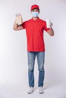 image d'un jeune livreur heureux en casquette rouge t-shirt blanc uniforme masque facial gants debout avec paquet de papier kraft marron vide isolé sur fond gris clair studio photo