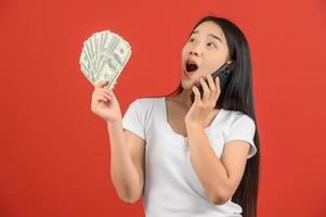 portrait d'une jeune femme joyeuse tenant des billets de banque et parlant sur un téléphone portable isolé sur fond rouge photo