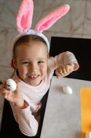 portrait d'une petite fille joyeuse, elle s'assied à la table et décore des oeufs de pâques, colle des autocollants avec des émotions dessus, dans ses mains se trouve un oeuf blanc qu'elle montre photo