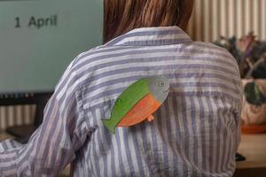 une jeune femme avec un poisson en papier par derrière à l'intérieur travaille sur un ordinateur sur lequel l'inscription le premier avril, poisson d'avril, blagues et blagues au bureau les unes sur les autres photo