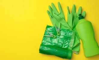 gants verts en caoutchouc pour le nettoyage, rouleau de sac en plastique poubelle et bouteille en plastique avec détergent sur fond jaune photo