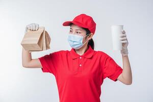 image d'un jeune livreur heureux en casquette rouge t-shirt blanc uniforme masque facial gants debout avec paquet de papier kraft marron vide isolé sur fond gris clair studio photo