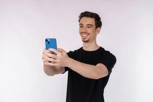 portrait d'heureux jeune bel homme en t-shirt noir tenant un téléphone et prenant une photo de selfie isolé sur fond blanc