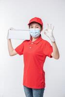 image d'un jeune livreur conscient en casquette rouge t-shirt blanc uniforme masque facial gants debout avec boîte en carton blanc vide isolé sur fond gris clair studio photo