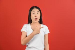 portrait d'une jeune femme asiatique excitée en t-shirt blanc pointant du doigt à côté isolé sur fond rouge photo