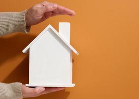 main féminine tient une maison en bois miniature blanche sur fond marron. concept d'assurance immobilière, protection de l'environnement, bonheur familial photo